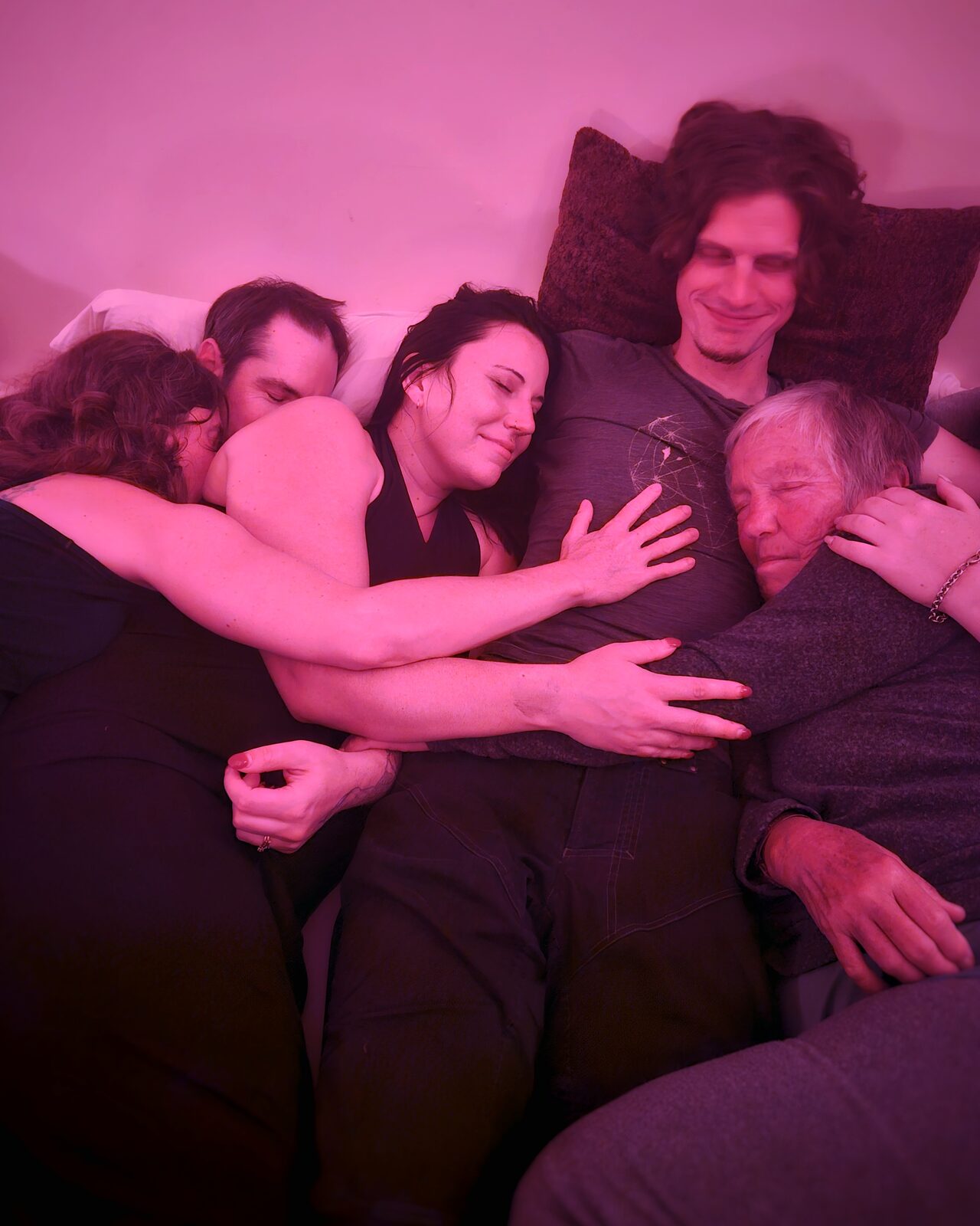 A close group cuddle under vivid pink light, with people resting peacefully against one another.