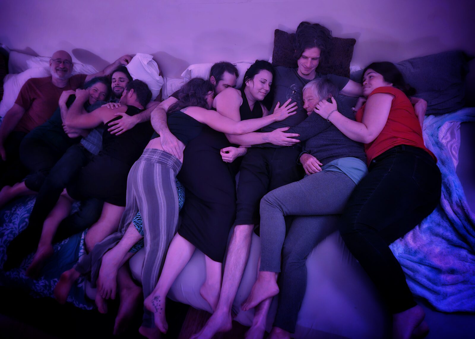 A large cuddle puddle of about ten people lying together on mattresses, lit in soft magenta.