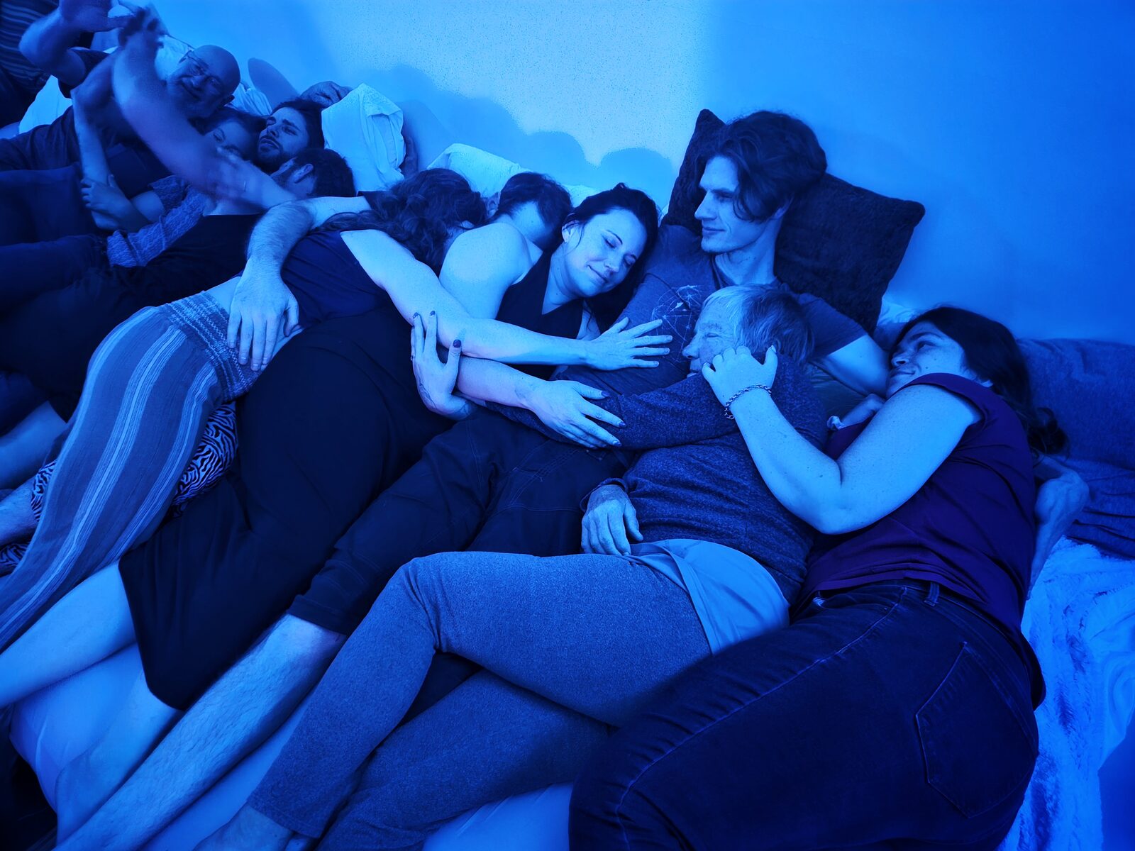 People nested closely together in a communal cuddle, the room washed in deep blue light.