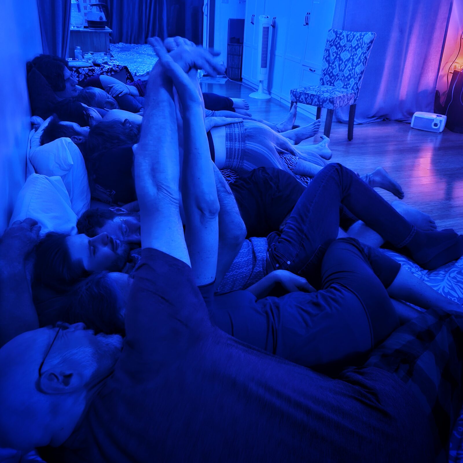 Arms reaching upward from a group resting together on the floor, under intense blue light.